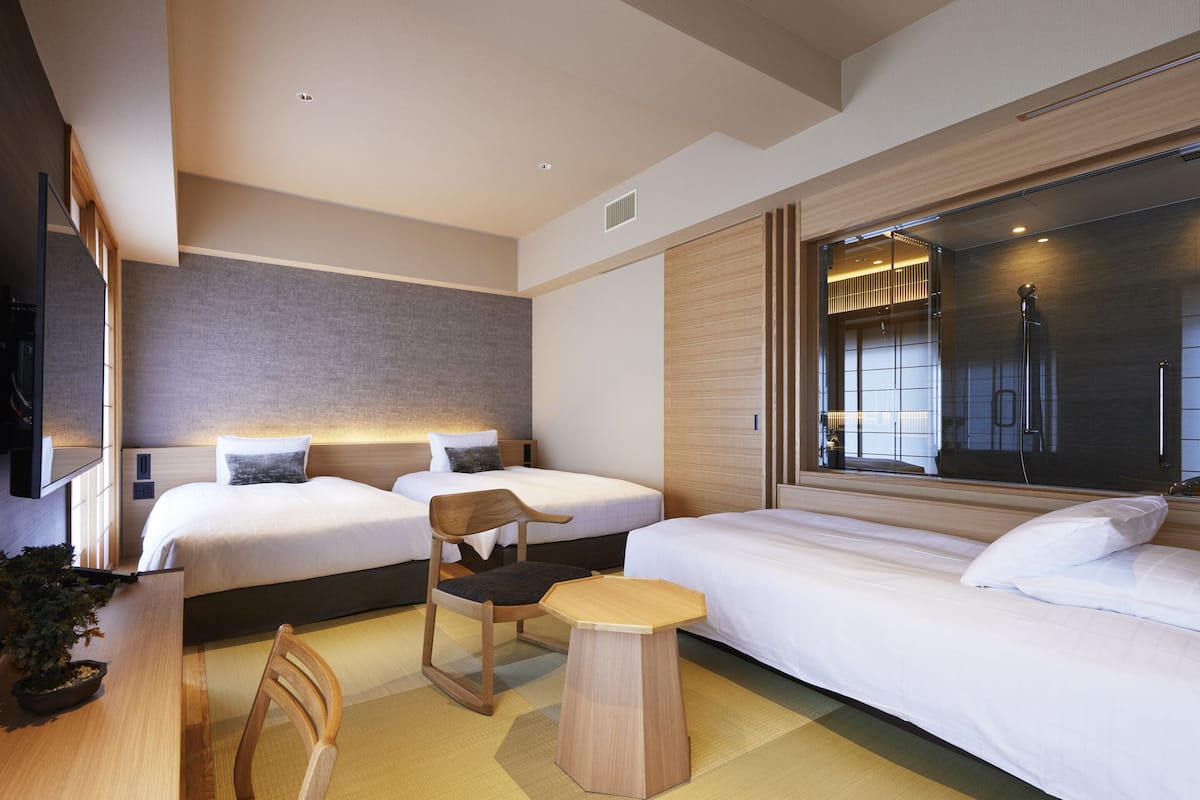 A modern hotel room with two beds, a wooden chair, a small table, and a large window with a view of the city.