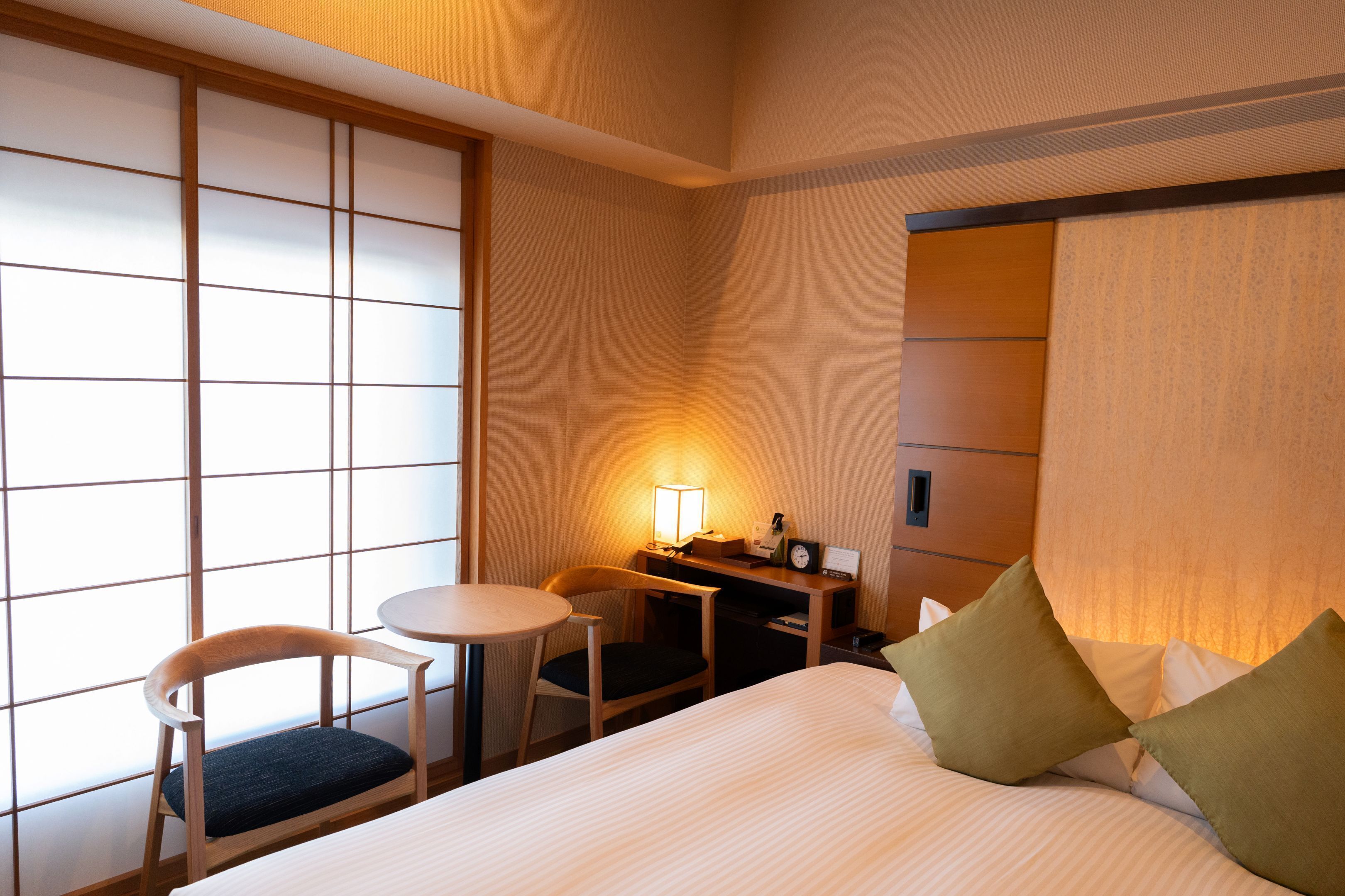 Habitación de hotel con cama, escritorio con lámpara, una silla y vista a través de un celosía japonesa.