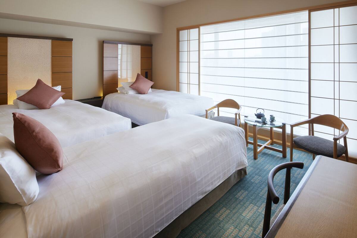 A hotel room with two beds, a small table with a chair, and a large window with blinds.