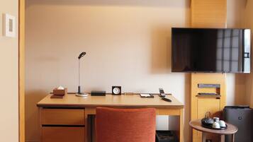 A hotel room with a wooden desk, a chair, a flat-screen TV, a small table with a kettle, and a trash bin.