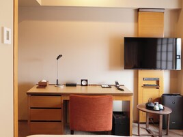 A hotel room with a wooden desk, a chair, a flat-screen TV, a small table with a kettle, and a trash bin.