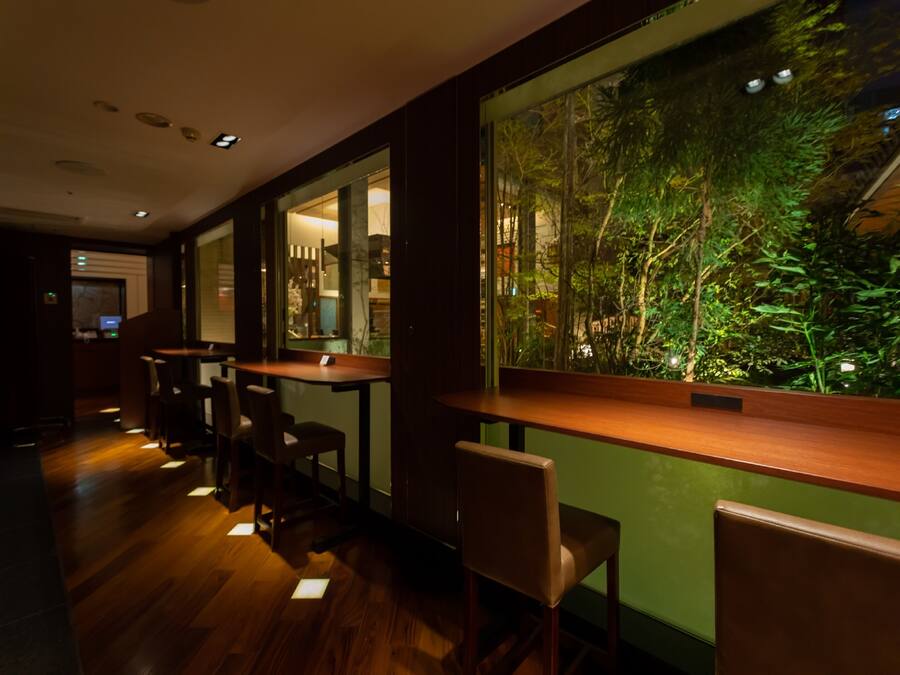 A dimly lit restaurant with wooden tables and chairs, a large window with a view of greenery, and a modern interior design.