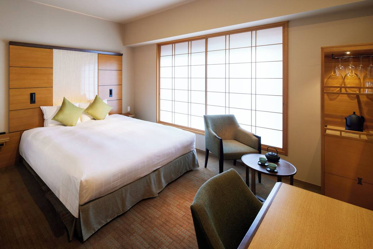 A hotel room with a large bed, a chair, a small table with tea set, and a wardrobe with hanging items.