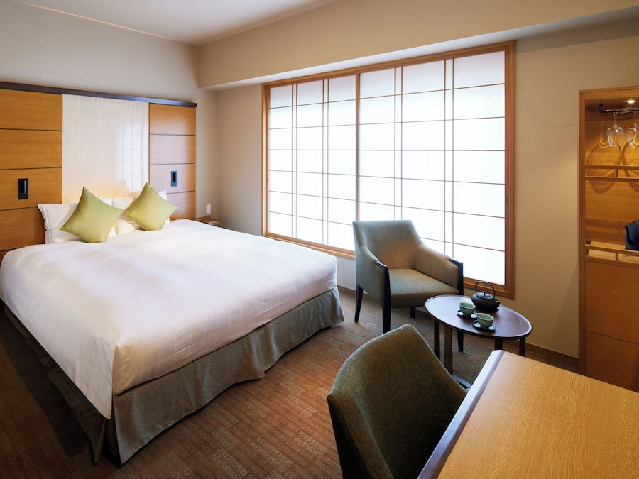 Quarto de hotel com uma cama grande, uma cadeira, uma mesinha com chá e um guarda-roupa cheio de roupas.