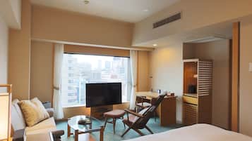 A hotel room with a bed, a TV, a desk, a chair, a sofa, and a window with a city view.