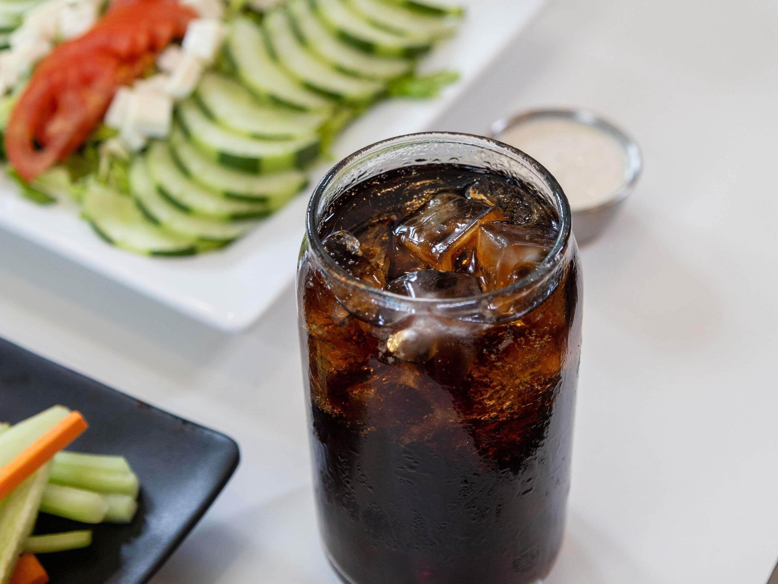 A glass of iced beverage with ice cubes, a plate of sliced vegetables, and a small bowl of sauce.