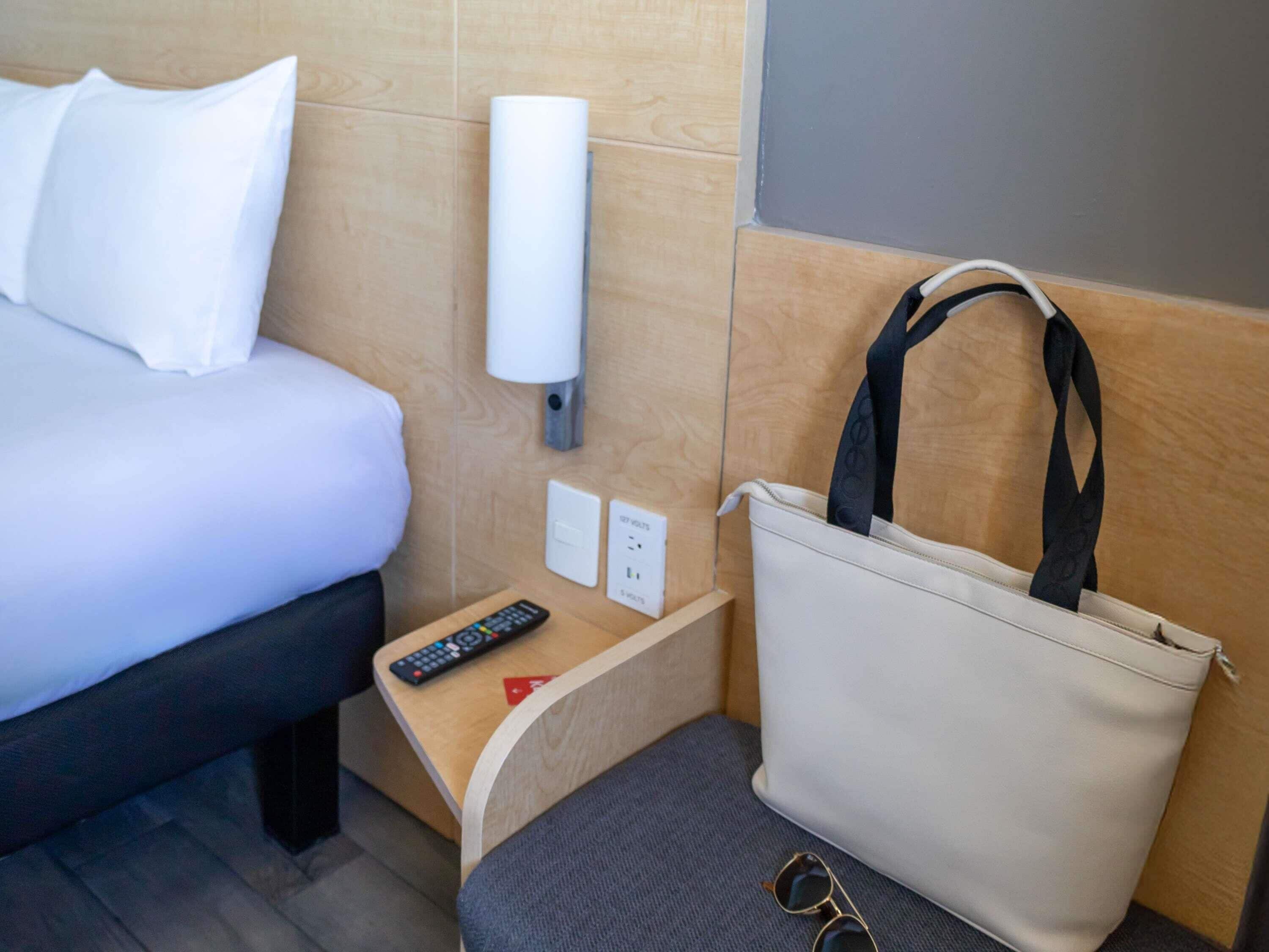 Une chambre d’hôtel avec un lit, une table de chevet avec une télécommande, une lampe fixée au mur et un grand sac à main posé sur le sol.