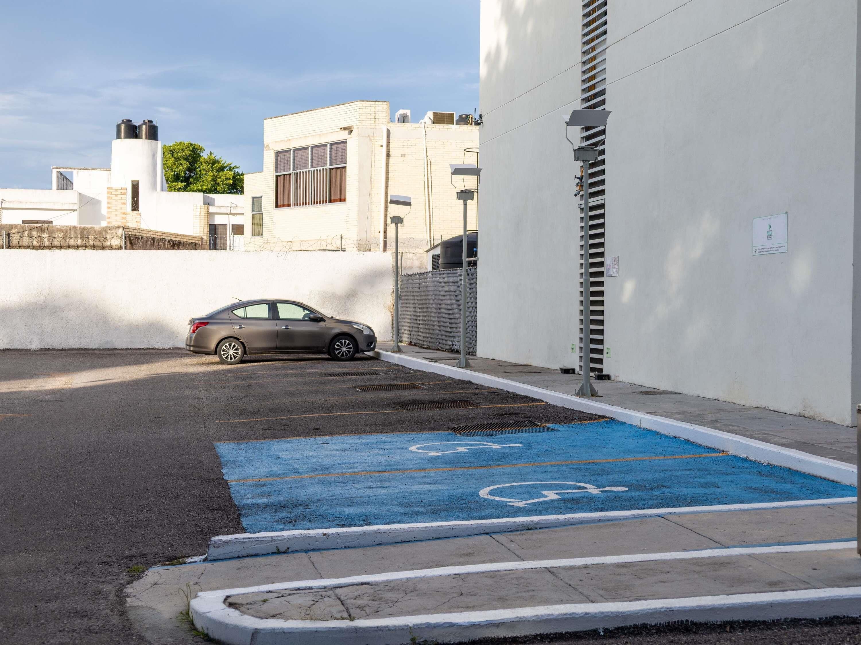 A parking area with a blue handicapped parking space, a car parked in the lot, and a building with a white facade and a barred fence.