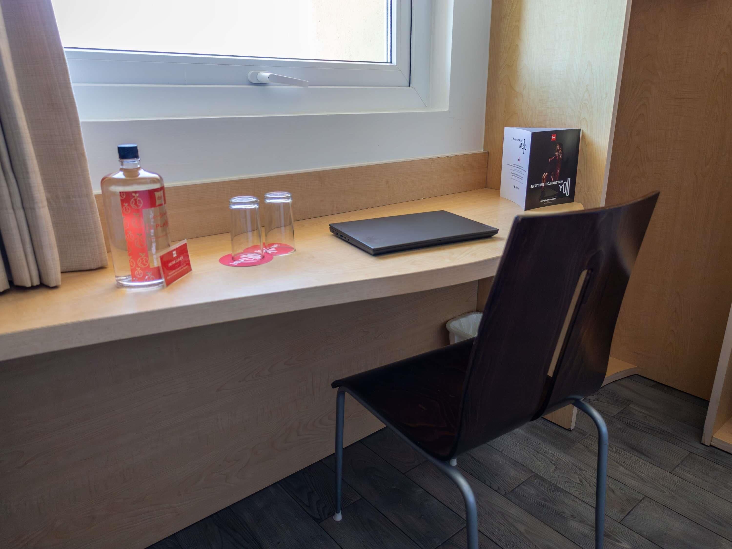 A hotel room with a wooden desk, a laptop, a chair, a bottle, and two glasses on the windowsill.