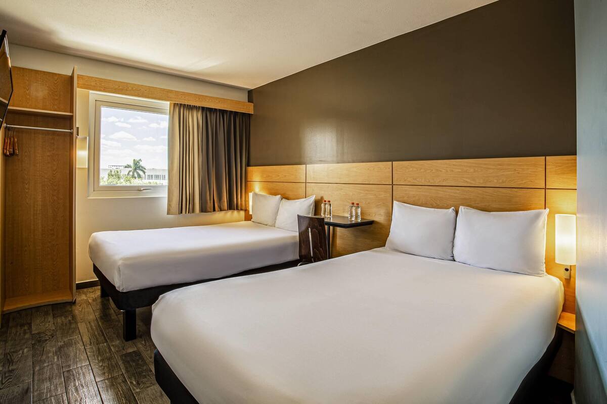 A hotel room with two beds, a window with curtains, a wooden headboard, and a small table with bottles.