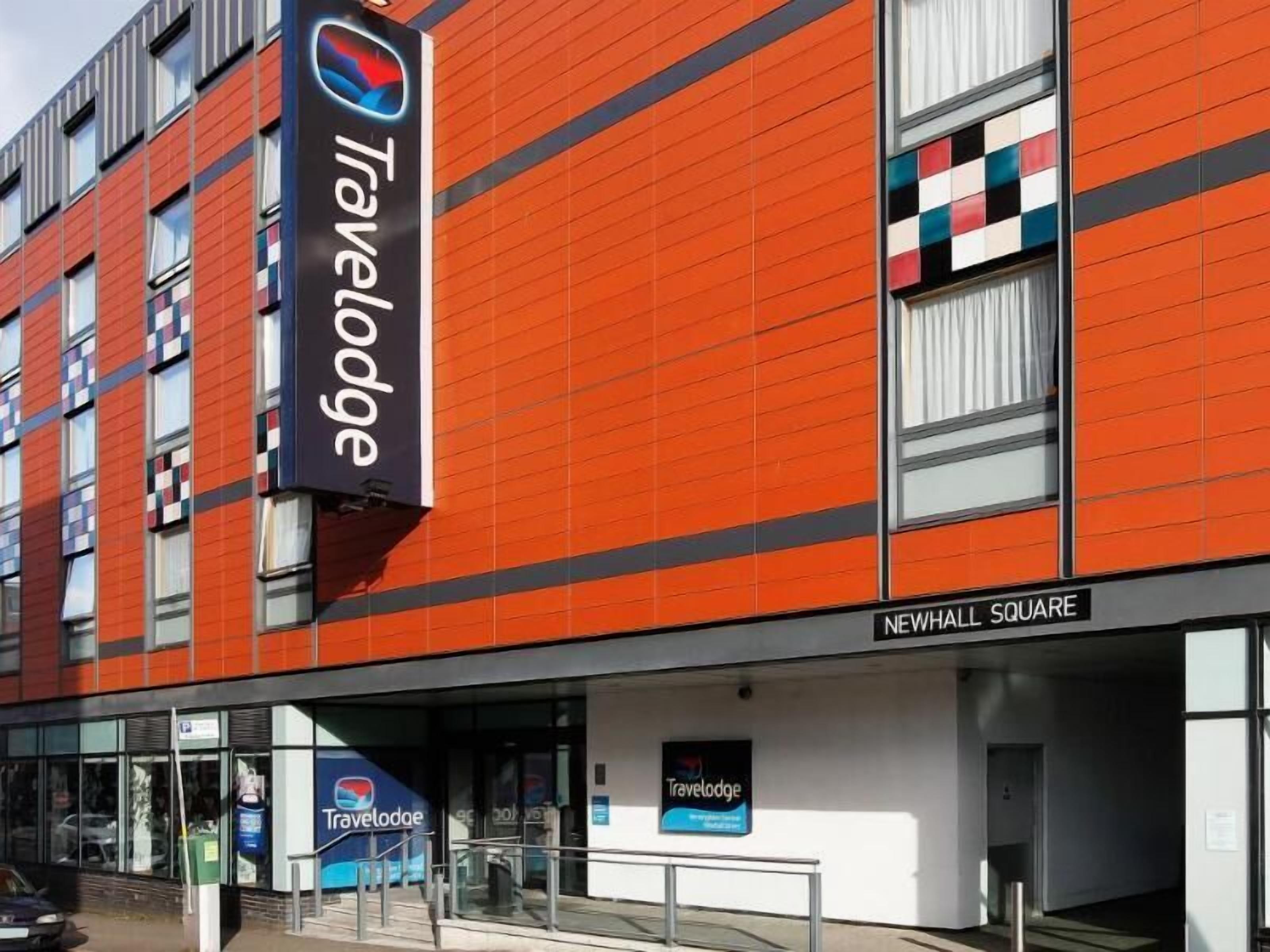 A Travelodge hotel with a modern orange facade, large windows, and a sign indicating Newhall Square.