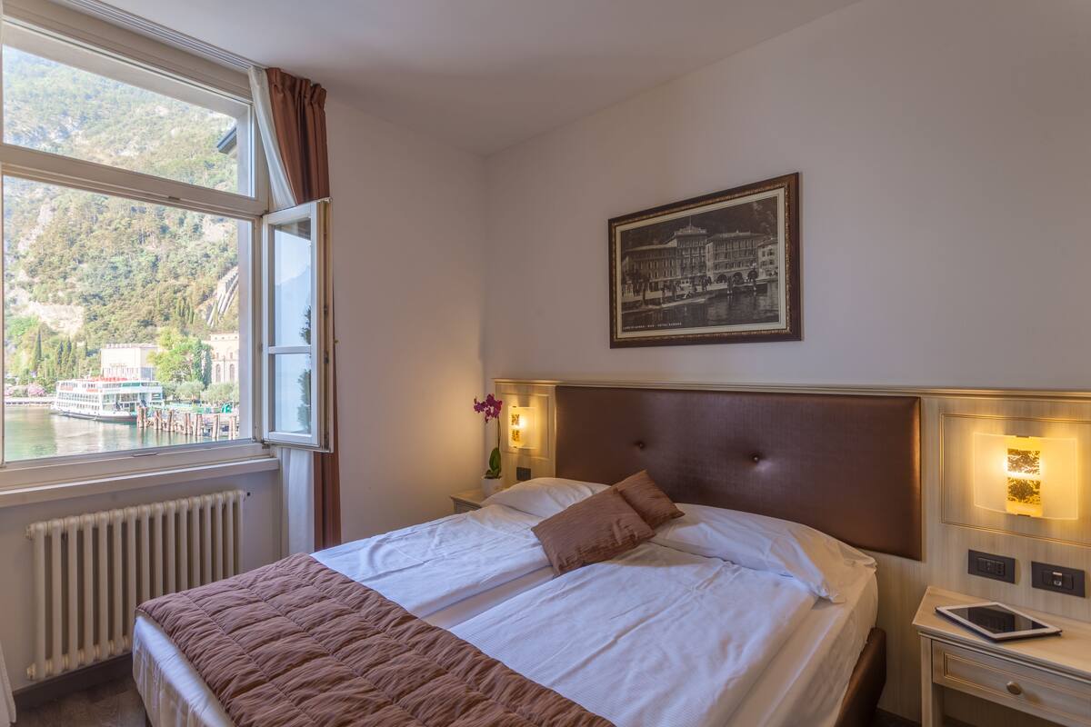 A hotel room with a large bed, a bedside table with a lamp, a framed picture on the wall, and a window with a view of a body of water and mountains.