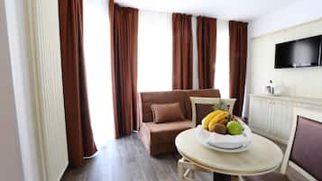 Ein modernes Hotelzimmer mit einem braunen Sofa, einem Esstisch mit Obstschale, einem Flachbildfernseher und Holzboden.