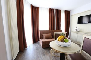 Una habitación de hotel moderna con un sofá marrón, una mesa de comedor con frutero, un televisor de pantalla plana y piso de madera.