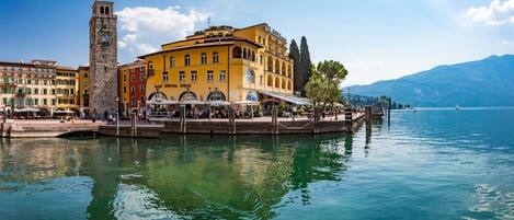 A waterfront hotel with outdoor seating, a clock tower, and a view of the lake and mountains.