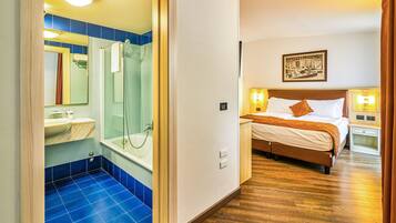 A hotel room with a bathroom featuring a bathtub, a shower, and a sink. The room has a bed with a wooden headboard and a nightstand with a lamp.