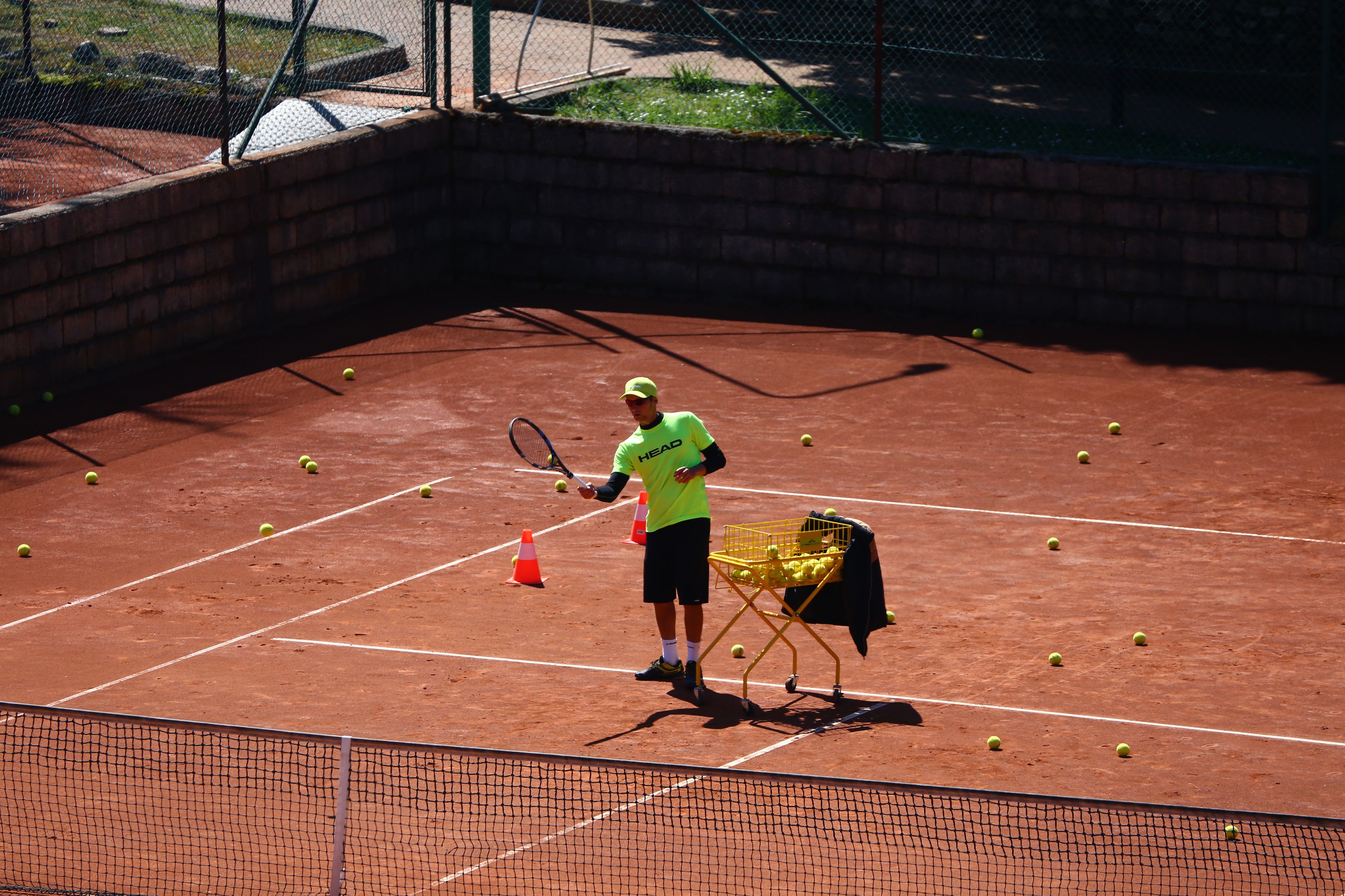 Una cancha de tenis con un jugador, una cesta de pelotas de tenis y una raqueta.