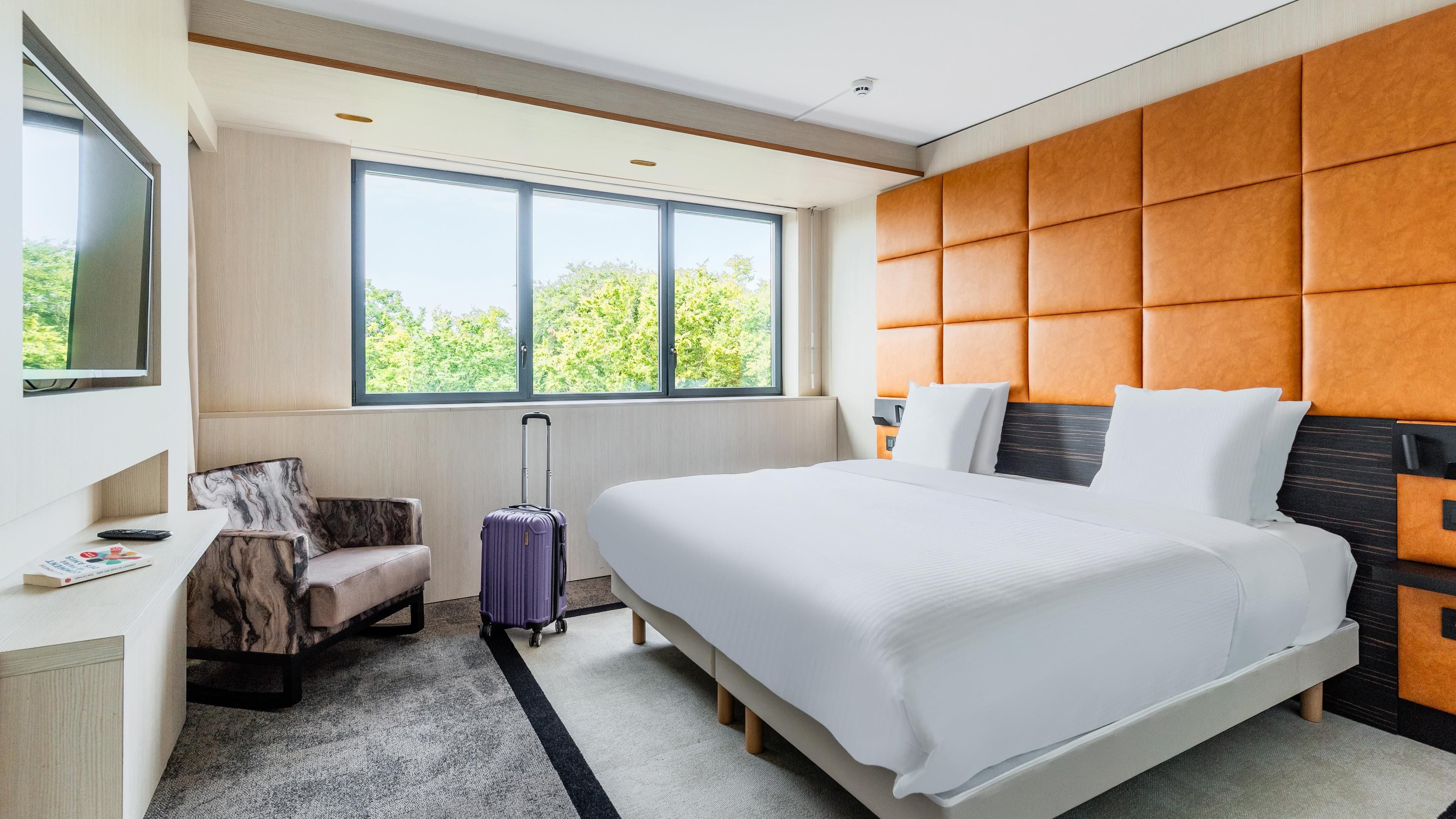Una habitación de hotel moderna con una cama grande, un área de descanso con una silla, una maleta y un ventanal que ofrece vistas a zonas verdes.