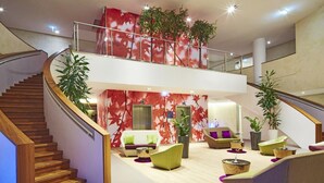 A modern hotel lobby with a wooden staircase, a seating area with sofas and chairs, and a red leaf-patterned wall.