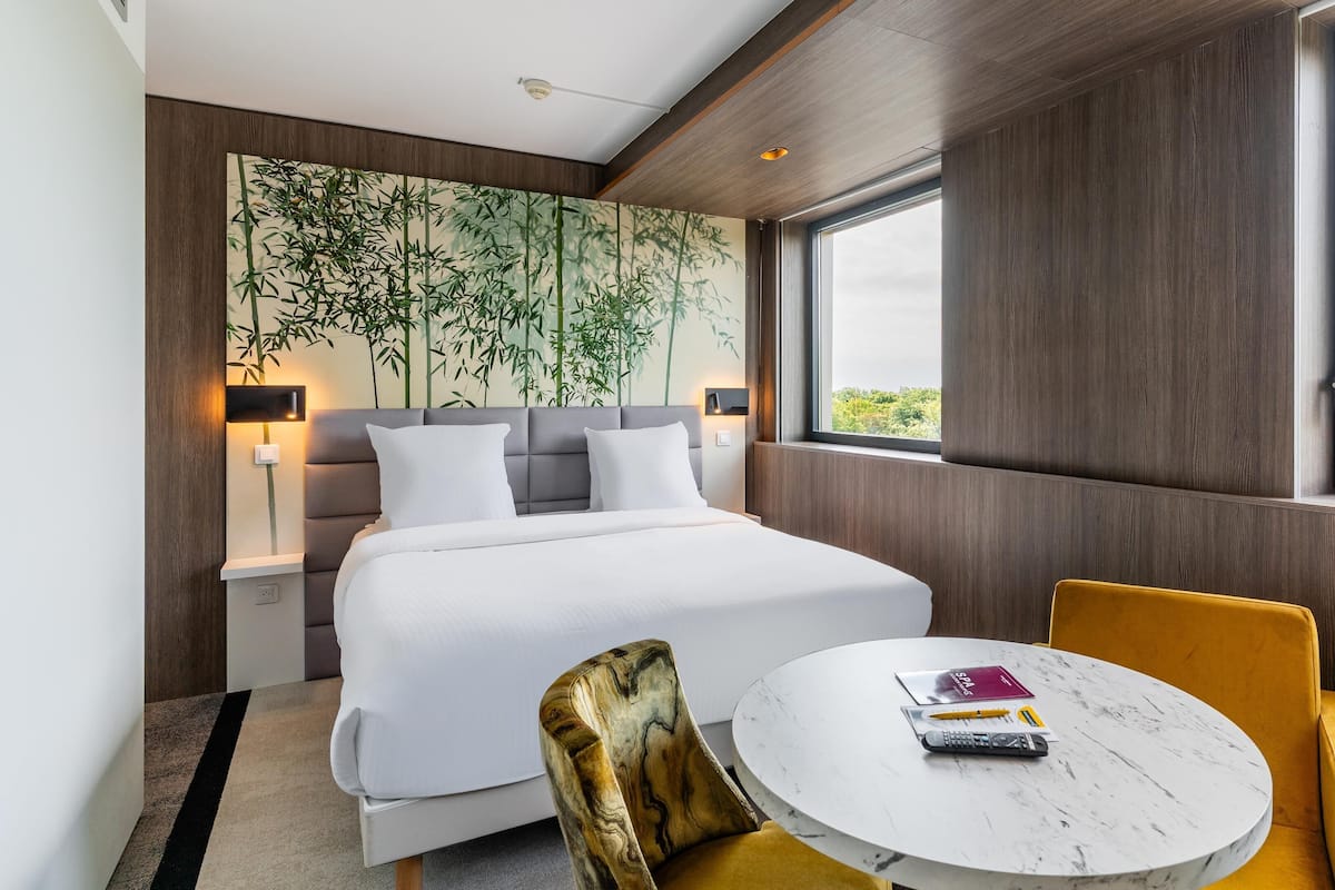 A modern hotel room with a large bed, a round table, and a view of greenery outside.