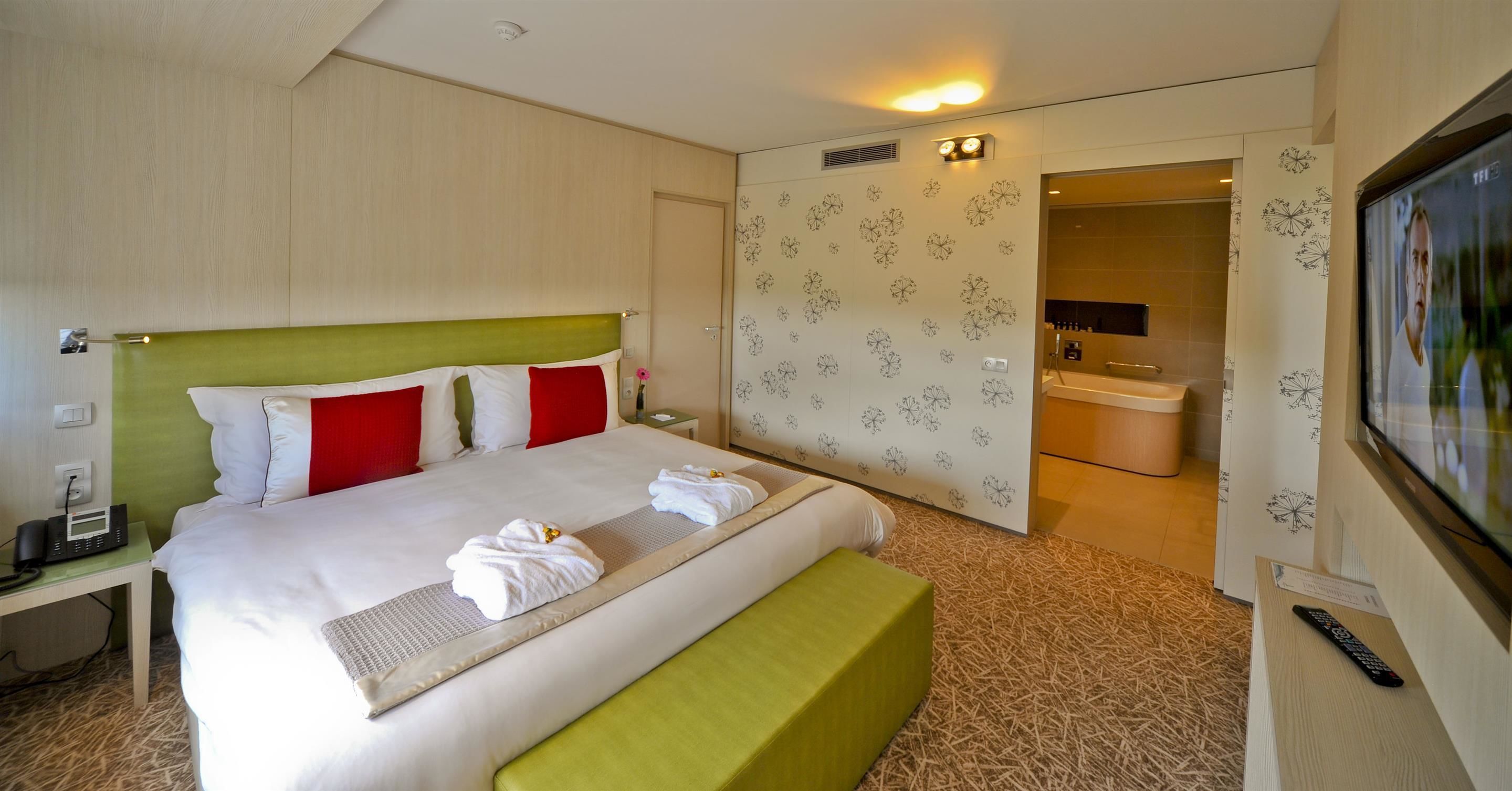 Une chambre d’hôtel avec un lit, un banc vert, une télévision et une salle de bain visible à travers une porte ouverte.