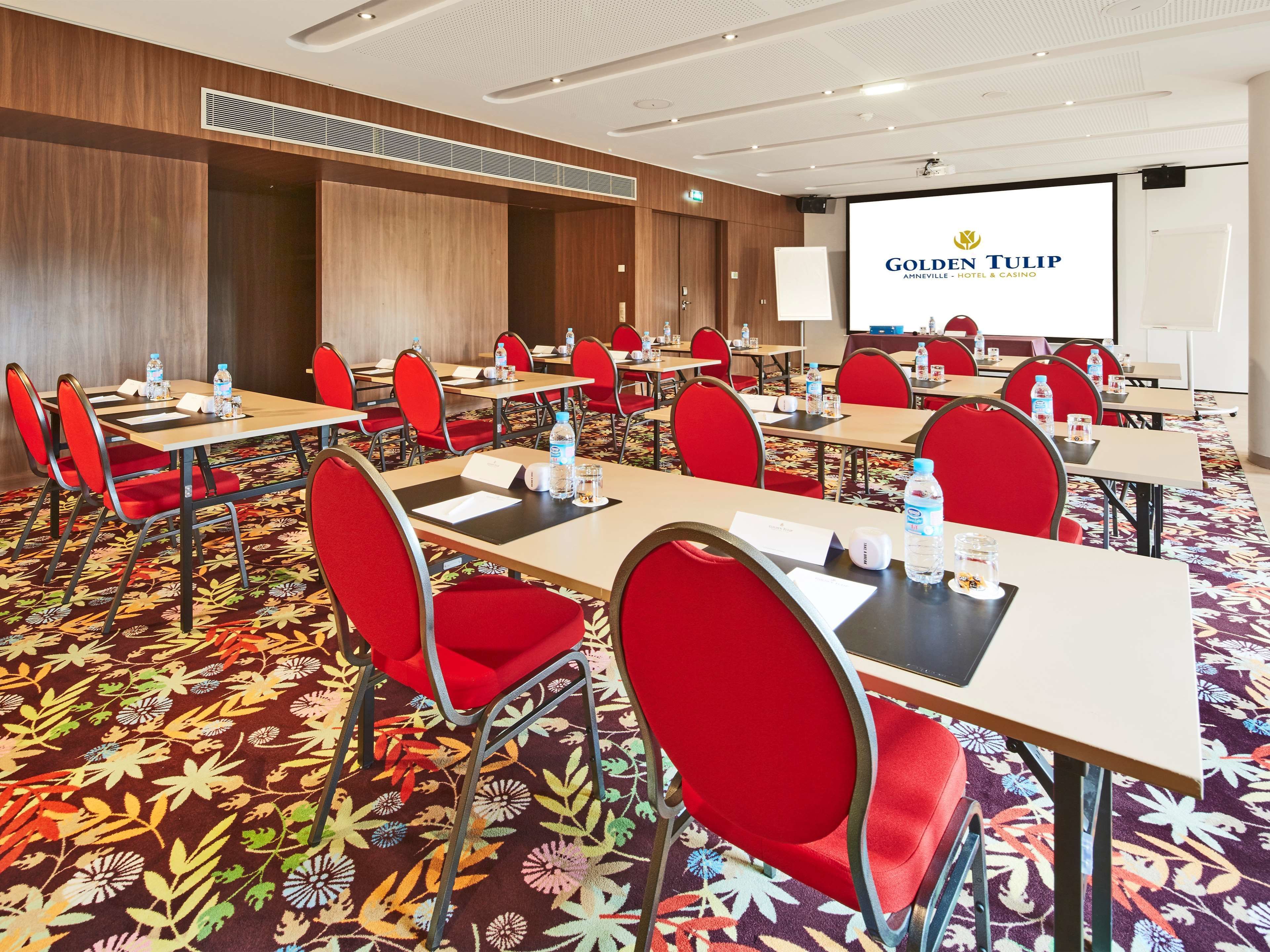 A conference room with red chairs and tables, a floral carpet, and a projector screen displaying "Golden Tulip Amneville Hotel & Casino".