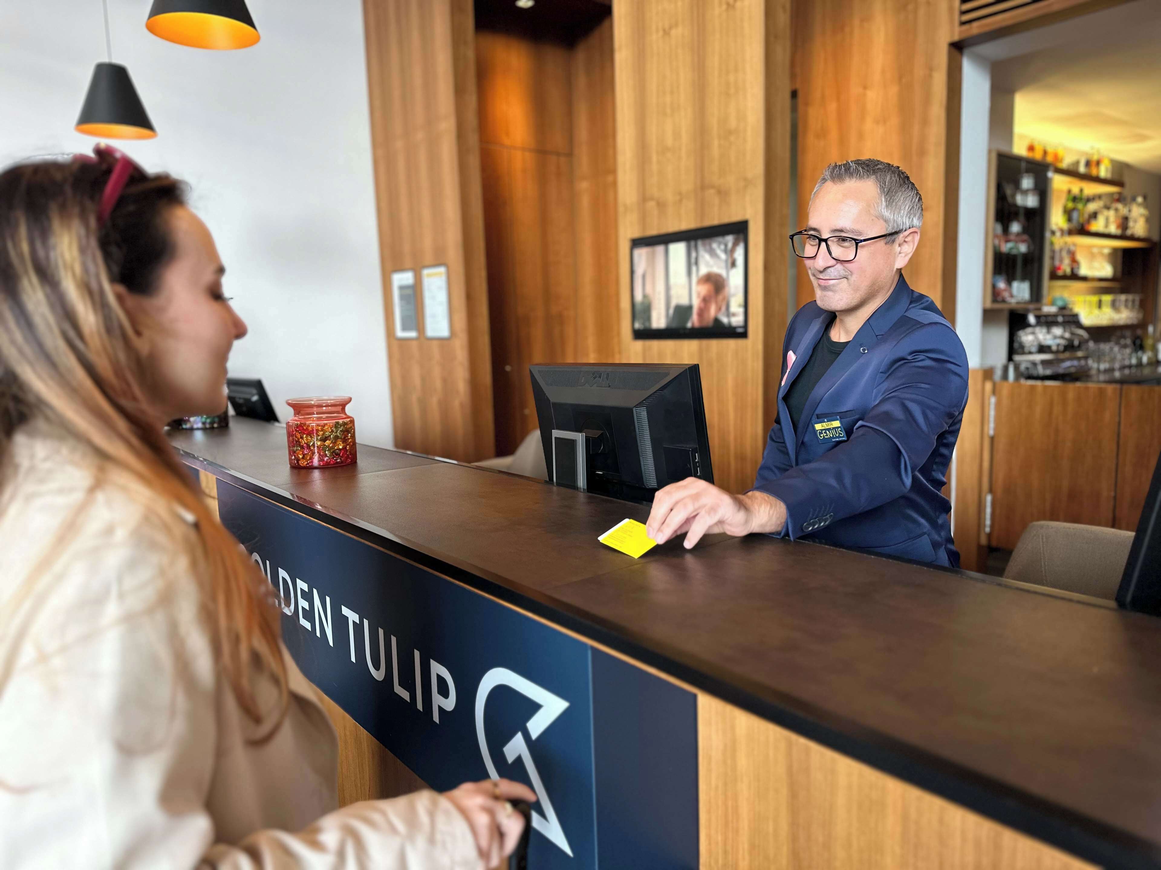 Un mostrador de recepción de hotel con interior de madera, una pantalla digital y un letrero que dice "DEN TULIP".