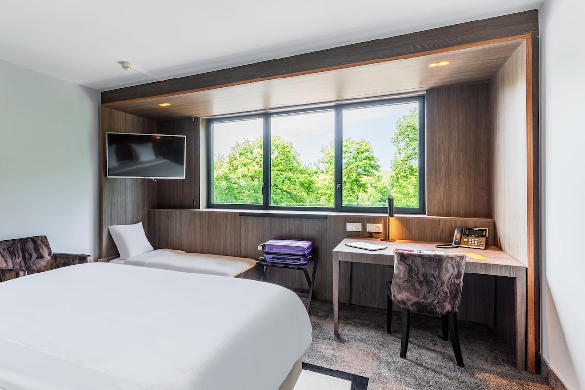 A hotel room with a large bed, a desk with a chair, a TV mounted on the wall, and a window with a view of trees.