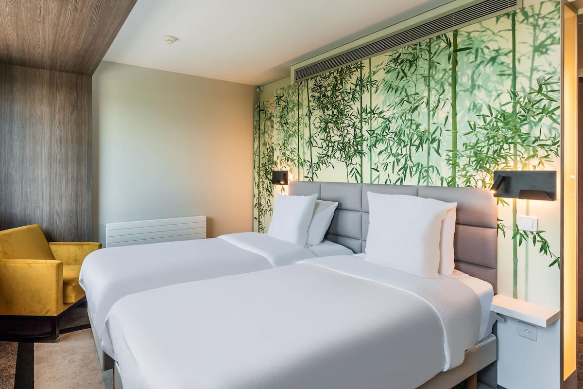 A hotel room with two beds, a yellow armchair, a wall with bamboo wallpaper, and a window with a view.