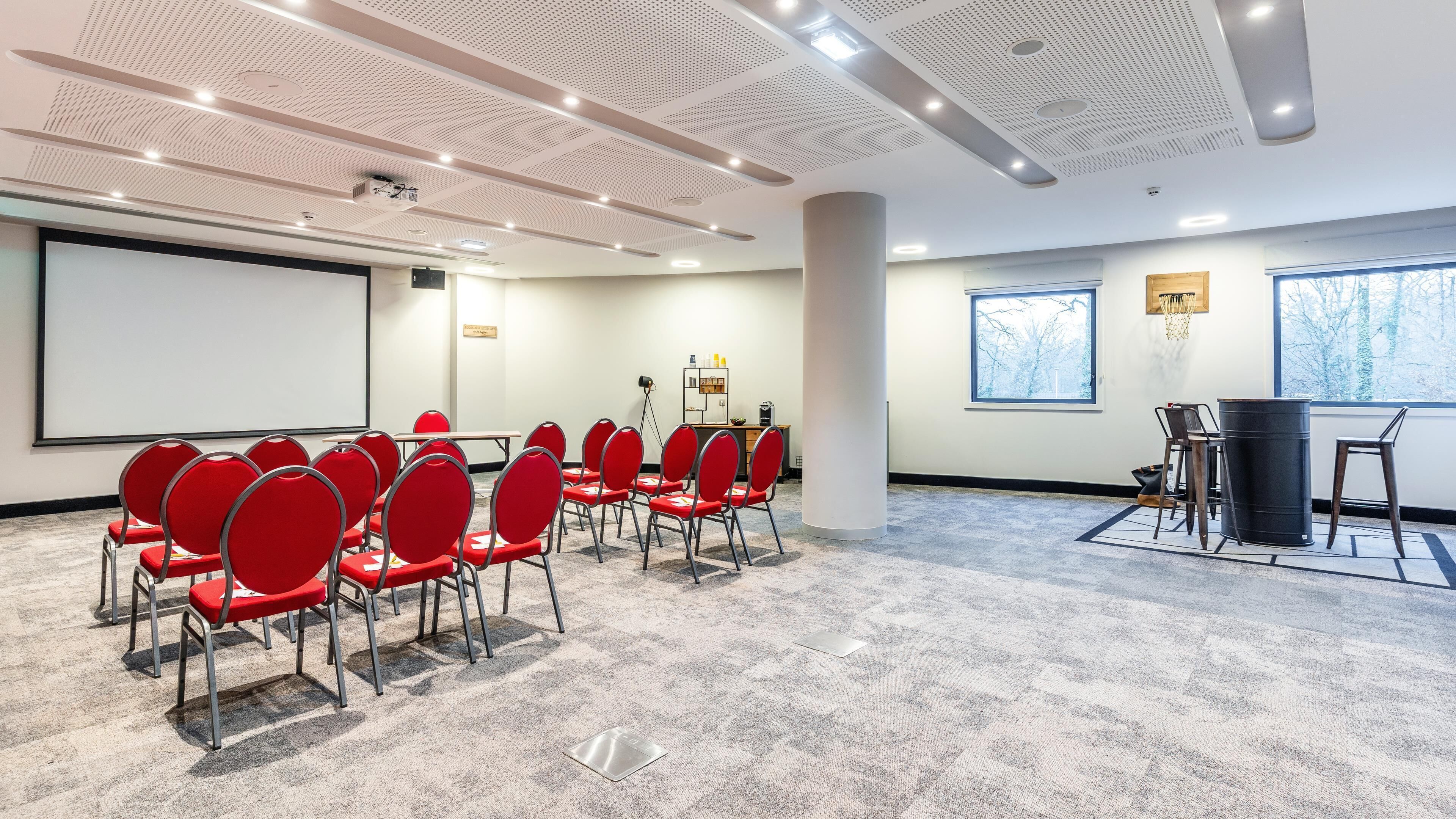Un salón de conferencias con sillas rojas, una pantalla de proyector, una pizarra y un amplio ventanal.