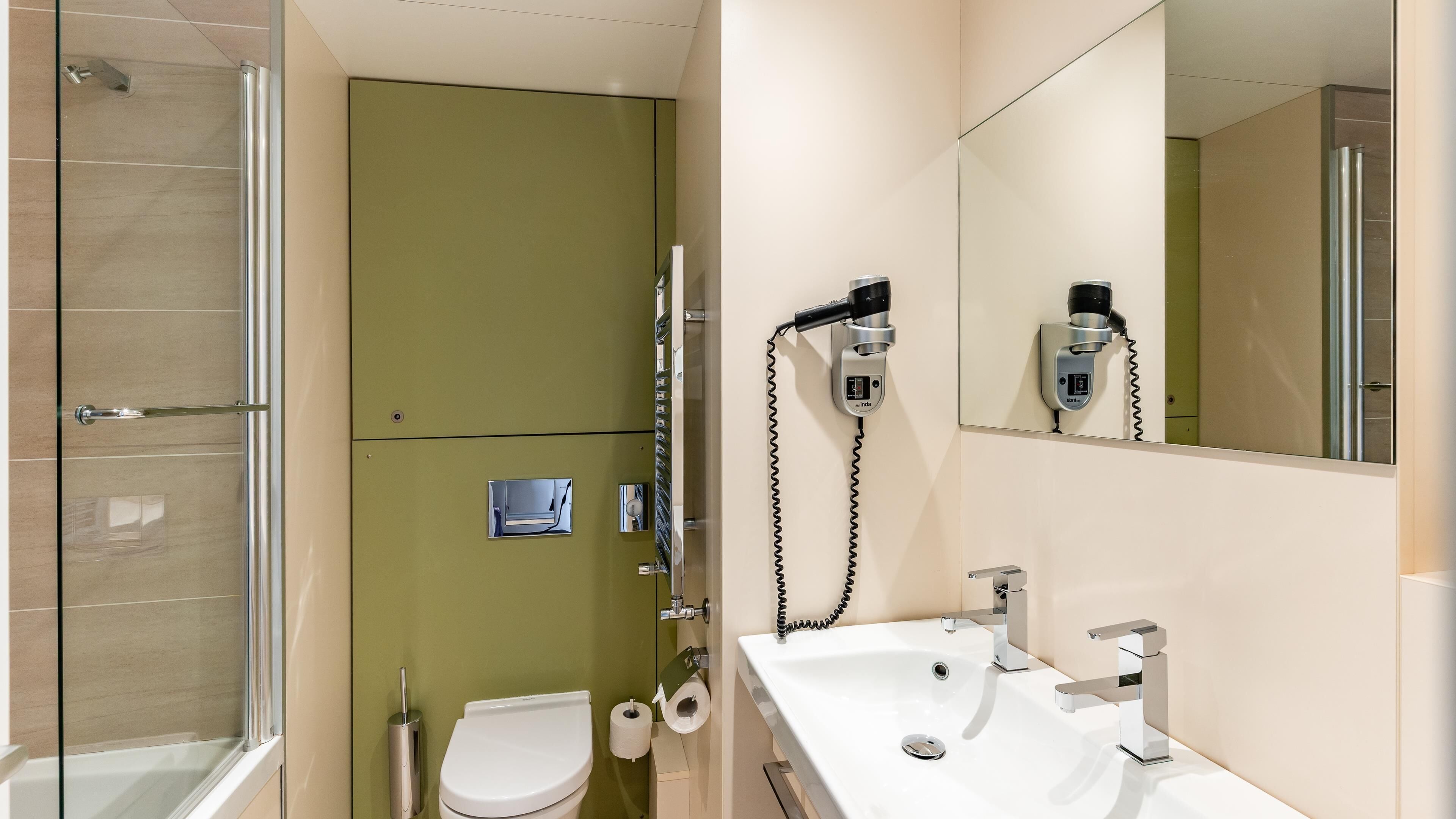 Un baño con cabina de ducha de vidrio, inodoro, lavabo con espejo y secador de pelo montado en la pared.