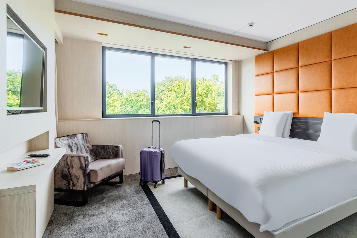 A modern hotel room with a large bed, a seating area with a chair, a suitcase, and a large window offering a view of greenery.