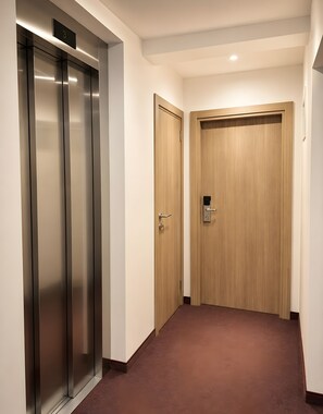 Un pasillo con un ascensor metálico, una puerta de madera con cerradura de llave electrónica y suelo alfombrado.