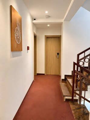 Un pasillo con una puerta de madera, un detector de incendios rojo y una escalera con barandilla metálica.