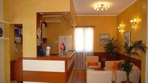 Zona de recepción de hotel con mostrador, área de espera con sillas y sofá, y cartel de recepción.