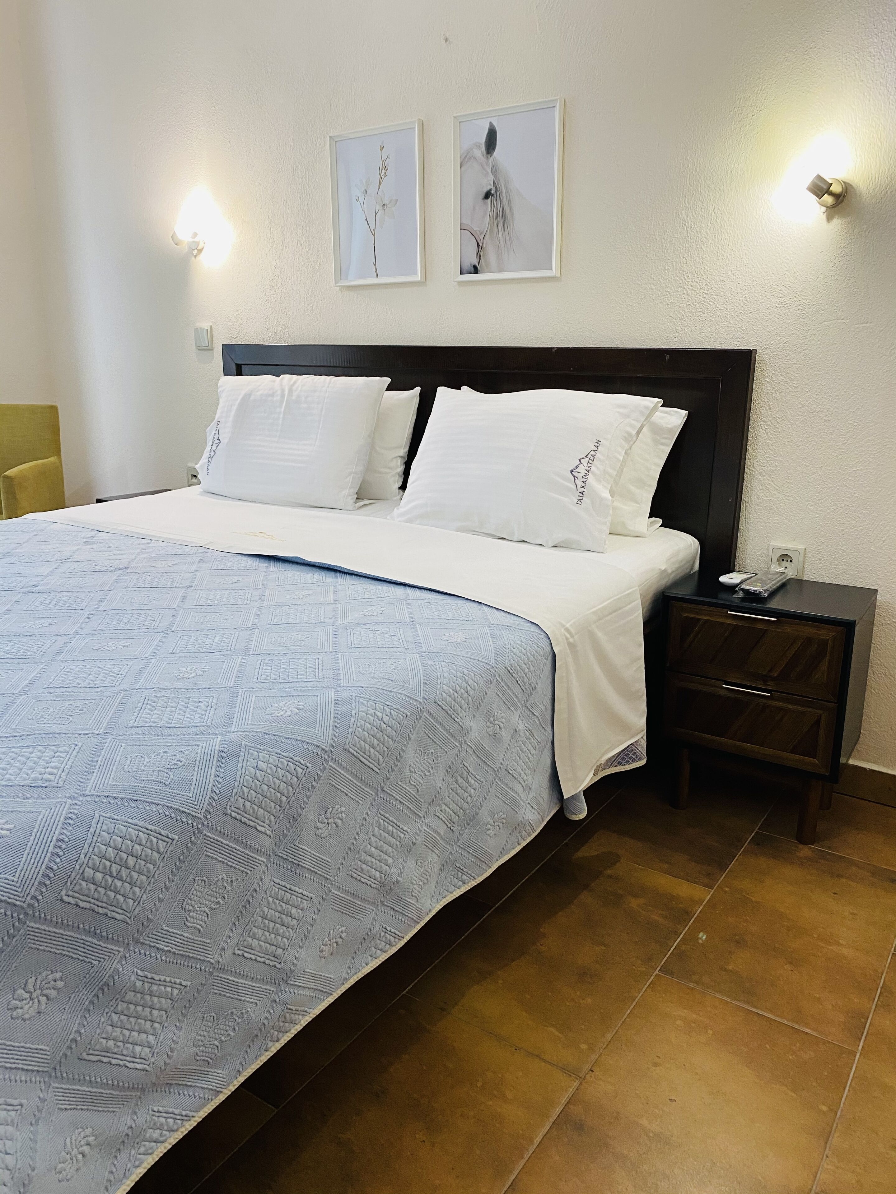 Una cama bien hecha con sábanas blancas y una colcha de cuadros, un cabecero de madera oscura y una mesita de noche con un control remoto.