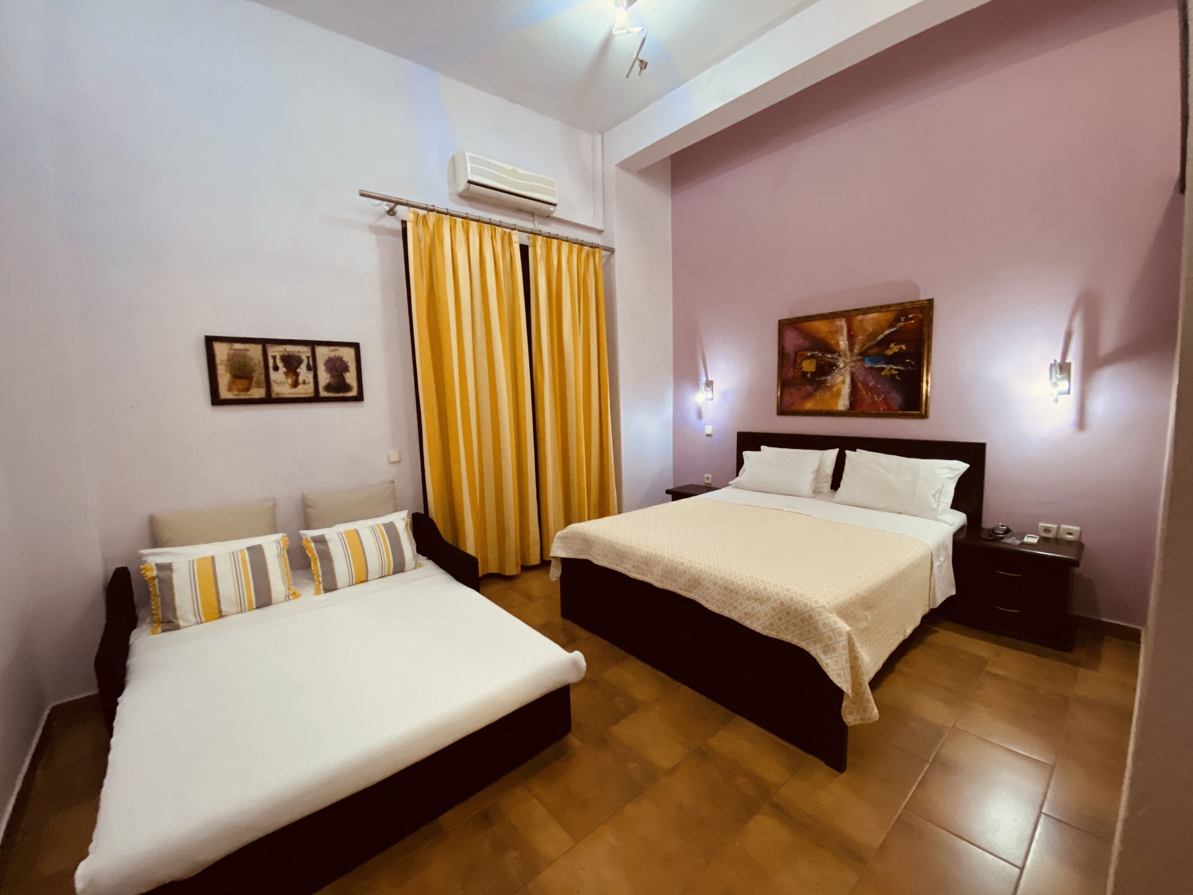 Una habitación de hotel con dos camas, un ventanal con cortinas amarillas, un aire acondicionado y un cuadro en la pared.
