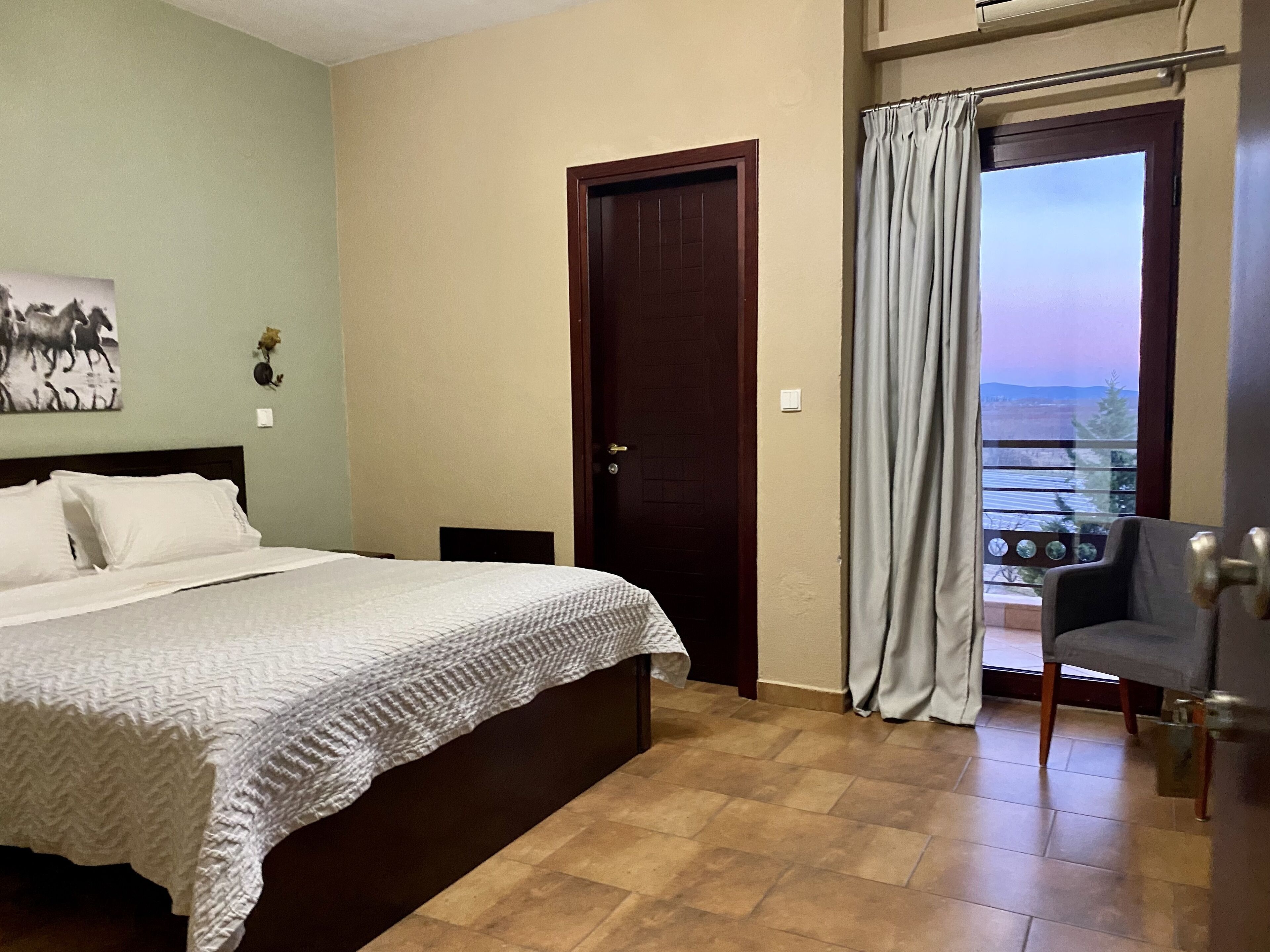 A bedroom with a bed, a door, a chair, and a balcony with curtains.