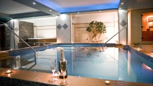 A modern indoor pool with a clear glass barrier, a lounge area with a potted plant, and a bar with a bottle and glasses on the poolside.