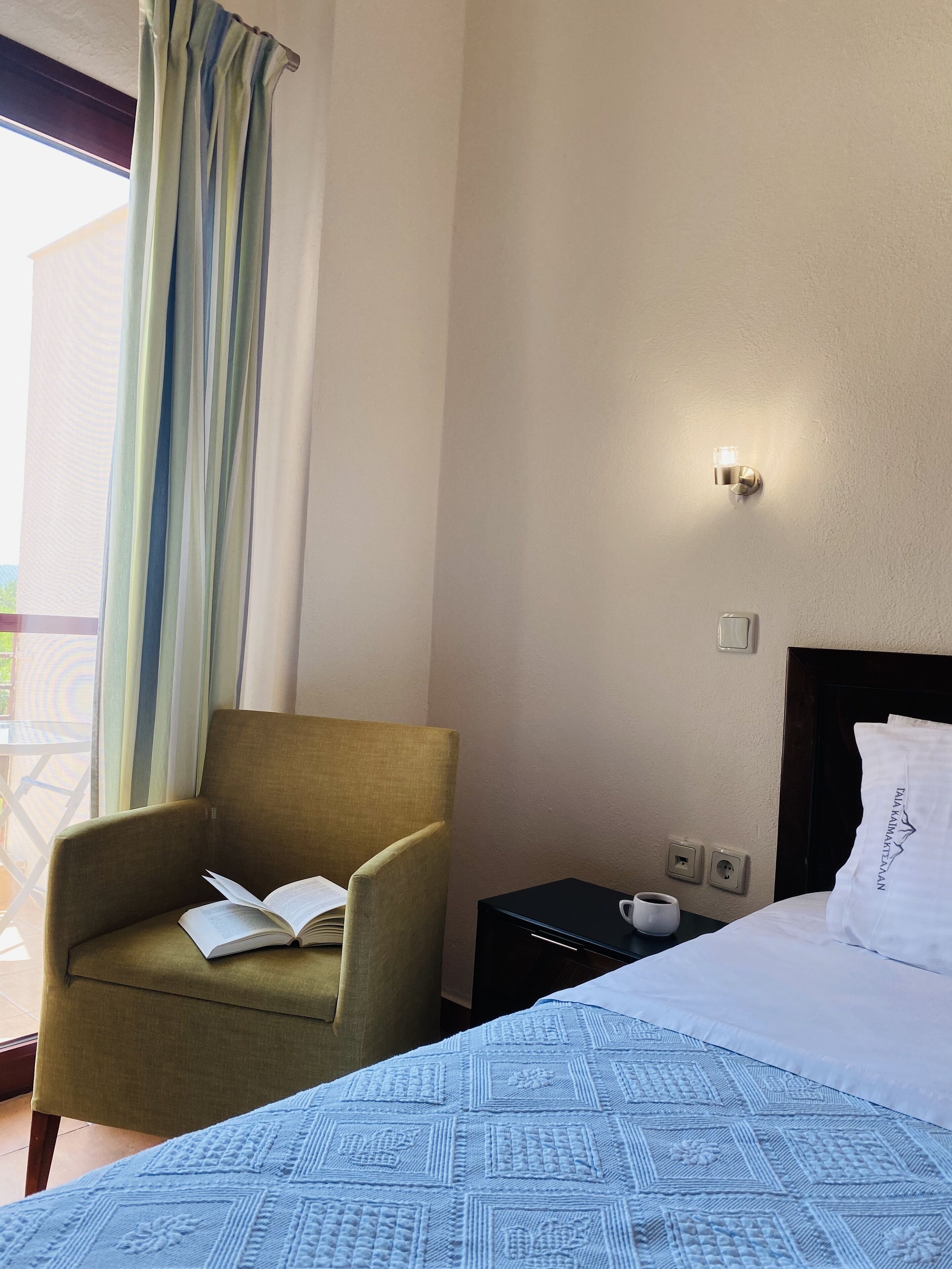 A hotel room with a bed, a green armchair with an open book, a bedside table with a cup, and a view of the outdoors through a window with curtains.