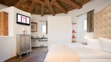A bedroom with a large bed, wooden ceiling beams, a vanity with a mirror, and a TV mounted on the wall.