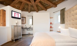 Une chambre dotée d’un grand lit, de poutres apparentes en bois au plafond, d’une coiffeuse avec un miroir et d’un téléviseur fixé au mur.