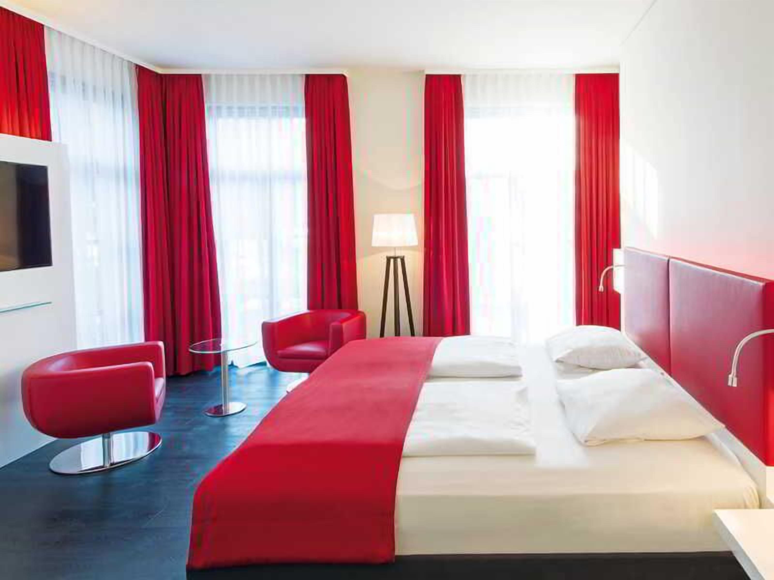 Een hotelkamer met een bed, twee rode stoelen, een vloerlamp en rode gordijnen.