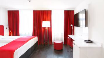 A hotel room with a bed, a desk, a chair, a red ottoman, and a TV mounted on the wall.