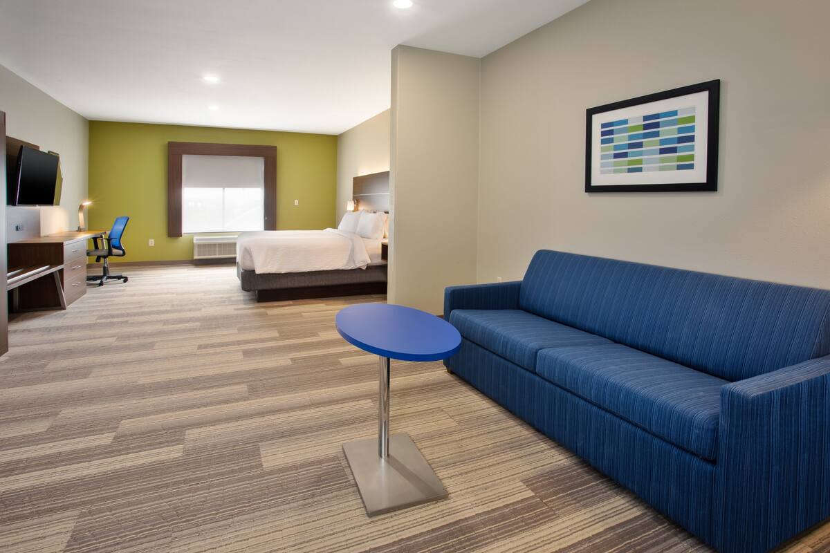 A hotel room with a bed, a blue sofa, a small round table, a desk with a chair, and a TV on the left.