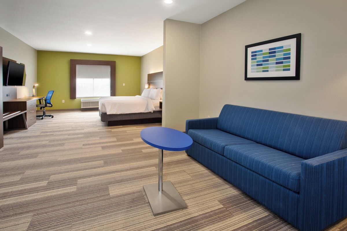 A hotel room with a bed, a blue sofa, a small round table, a desk with a chair, and a TV on the left.