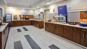 A modern kitchen area with wooden cabinets, a digital menu board, and a variety of food items displayed on the counter. - Holiday Inn Express & Suites Houston West - Katy by IHG (Katy)