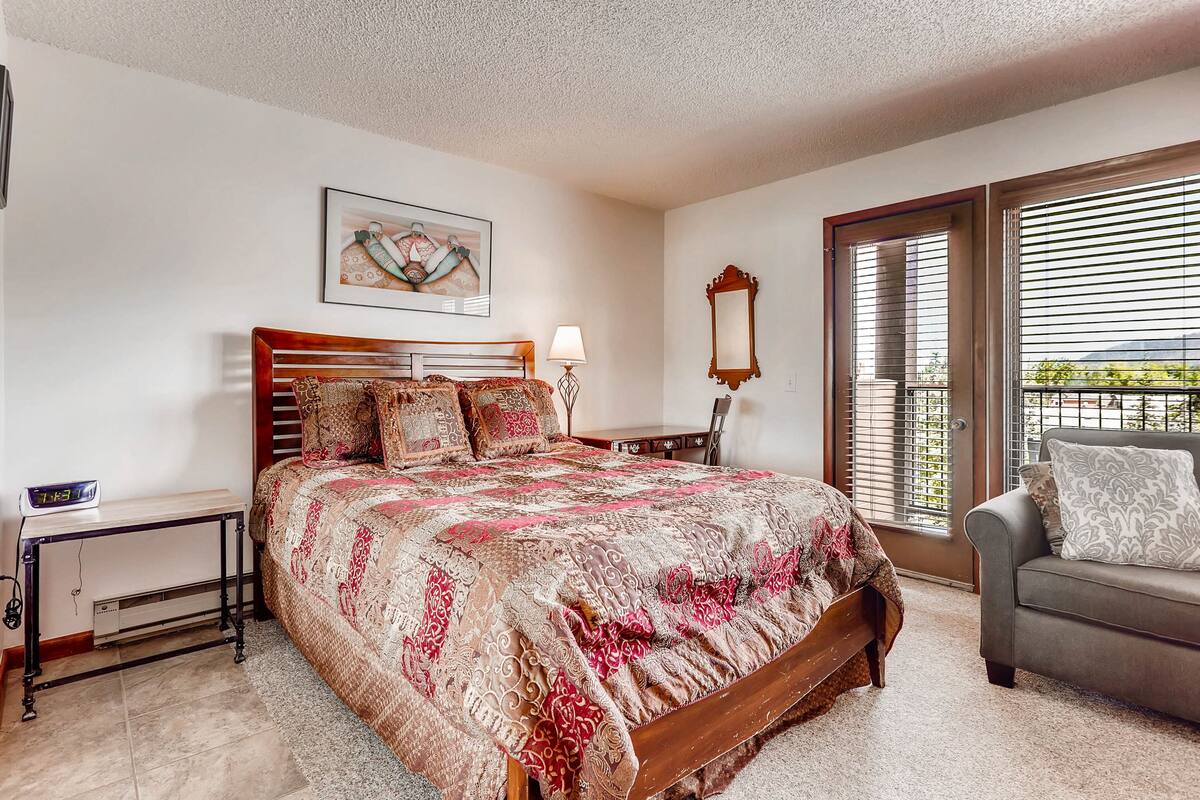 A bedroom with a large bed, a bedside table, a lamp, a chair, a window with blinds, and a painting on the wall.