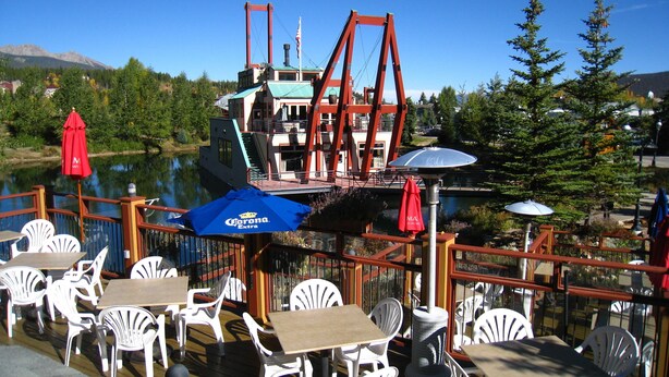 Un área de comedor al aire libre con mesas y sillas, un barco en el fondo y vistas a un lago y montañas.