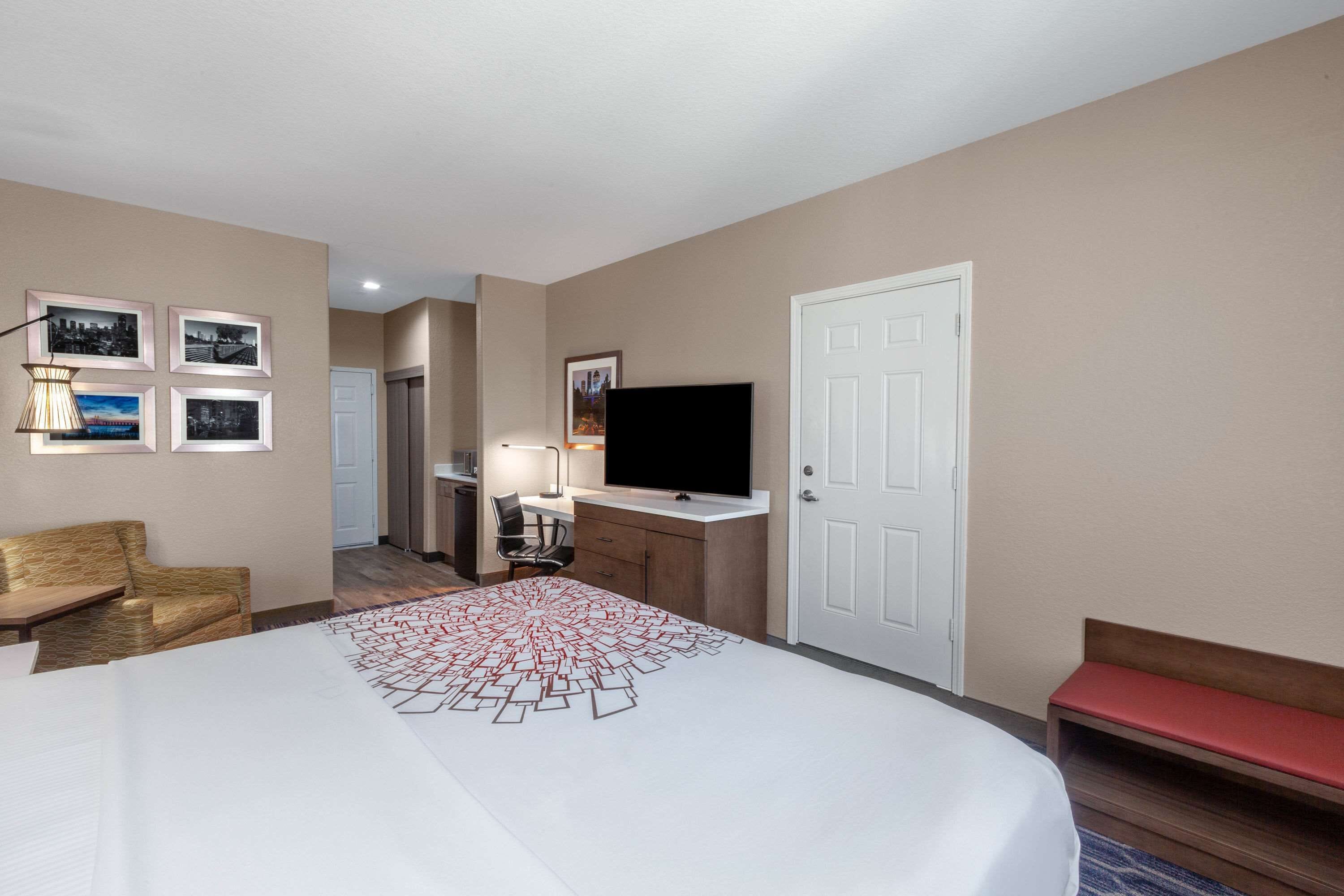 A hotel room with a large bed, a desk with a flat-screen TV, a chair, a lamp, and a door.